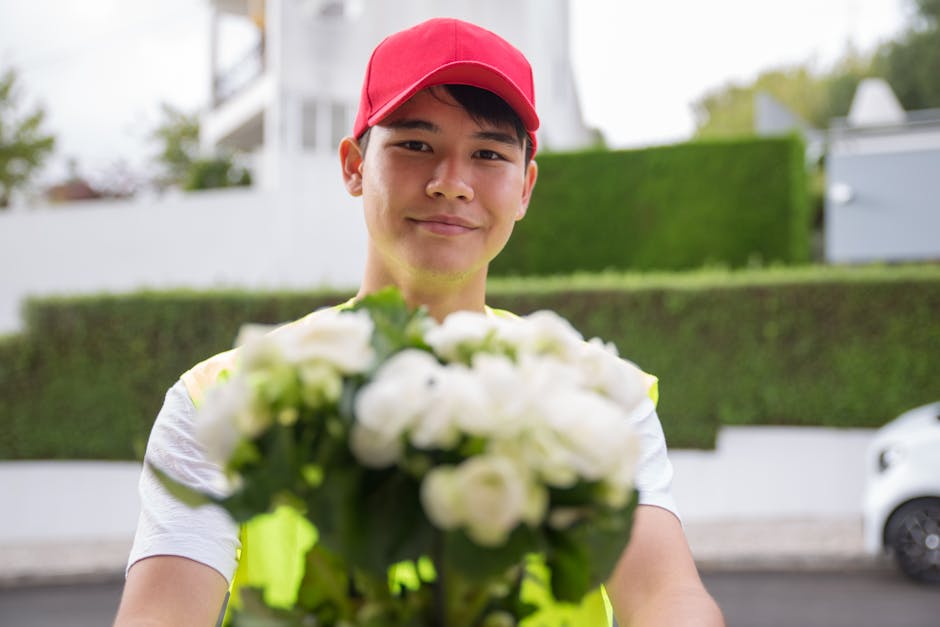 A delivery person wearing a red cap and high-visibility vest presents a bouquet of fresh white roses and green foliage, arranged in a compact floral gift. The bouquet is wrapped in clear cellophane, s