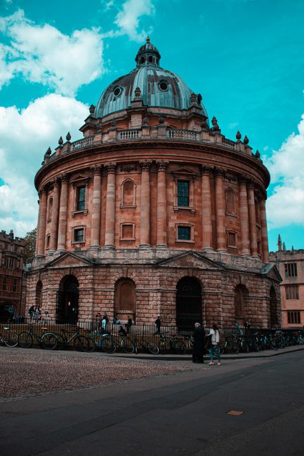 A photograph of the Radcliffe Camera in Oxford showing its distinctive circular architecture with a large, domed roof topped by a small lantern and cupola. The building's exterior is made of warm redd