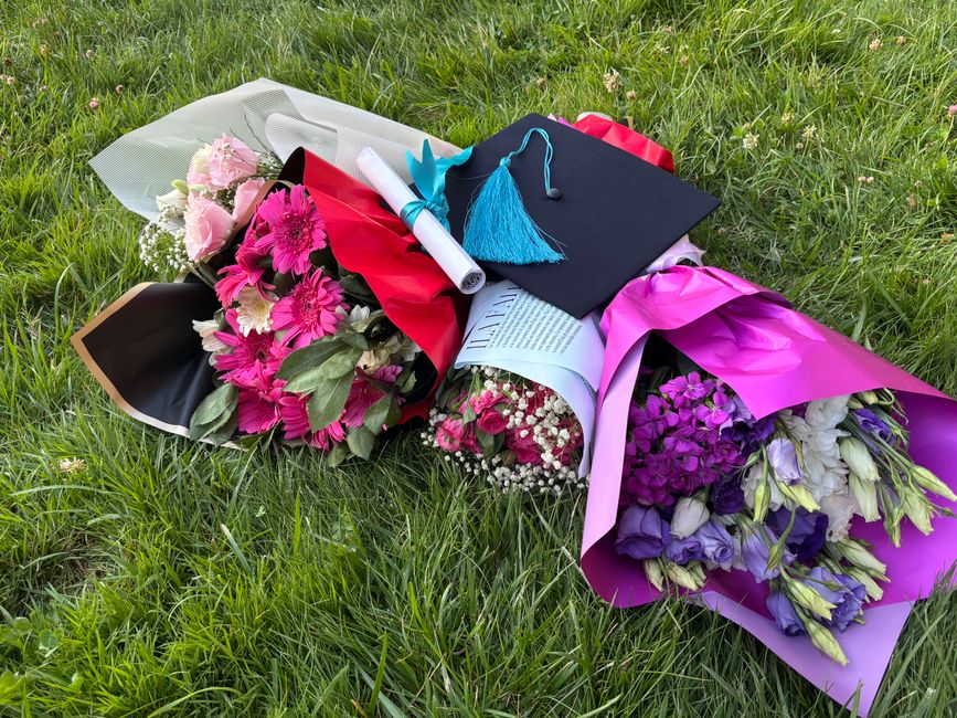A collection of vibrant flower bouquets lies on lush green grass, featuring predominantly pink, purple, and white flowers such as gerberas, irises, and roses, wrapped in colorful paper including black