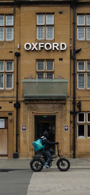 The image depicts the exterior of a historic sandstone building with the word 'OXFORD' displayed prominently in white capital letters above the entrance. In front of the building, a delivery person dr
