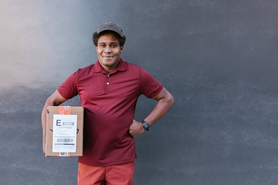 A delivery person with dark skin, short curly hair, wearing a maroon polo shirt, red pants, and a grey cap, standing against a plain grey wall. He is smiling and holding a small cardboard parcel with 